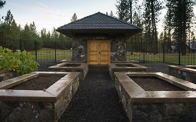 A small stone building with wooden double doors stands in the center, surrounded by four empty raised garden beds bordered with stone—showcasing Copper Creek Landscaping’s attention to detail. Pine trees and grass are visible in the background.
