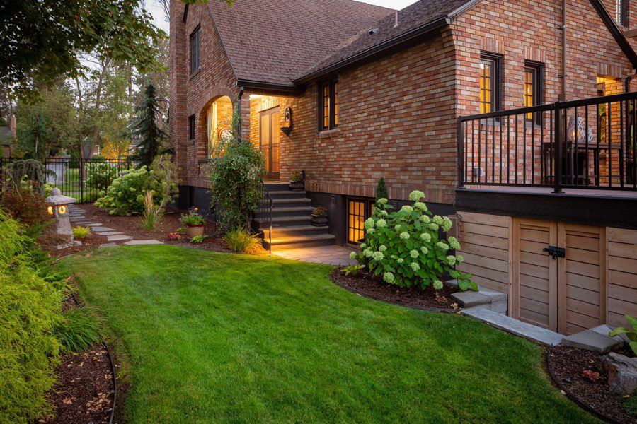 A brick house with illuminated windows, a small porch with stairs, Copper Creek Landscaping features a landscaped garden, green lawn, shrubs, and stone path. There is a gated fence on the left and doors leading to a basement under a raised deck.