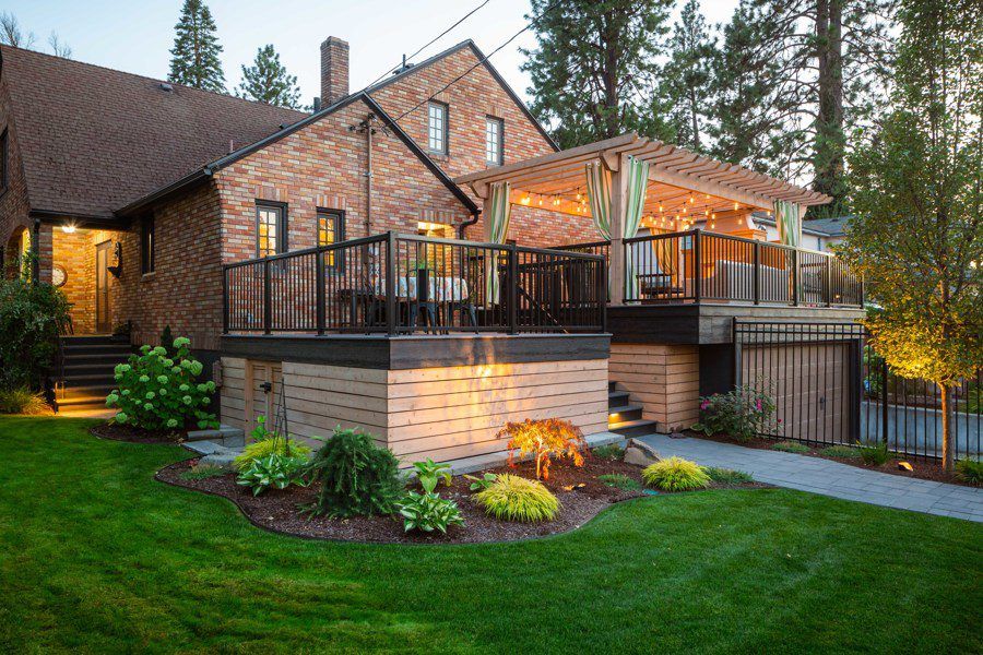 A brick house with a large wooden deck featuring outdoor dining and lounge areas under a pergola with string lights, surrounded by professionally designed Copper Creek Landscaping, including a well-maintained lawn, shrubs, and trees.