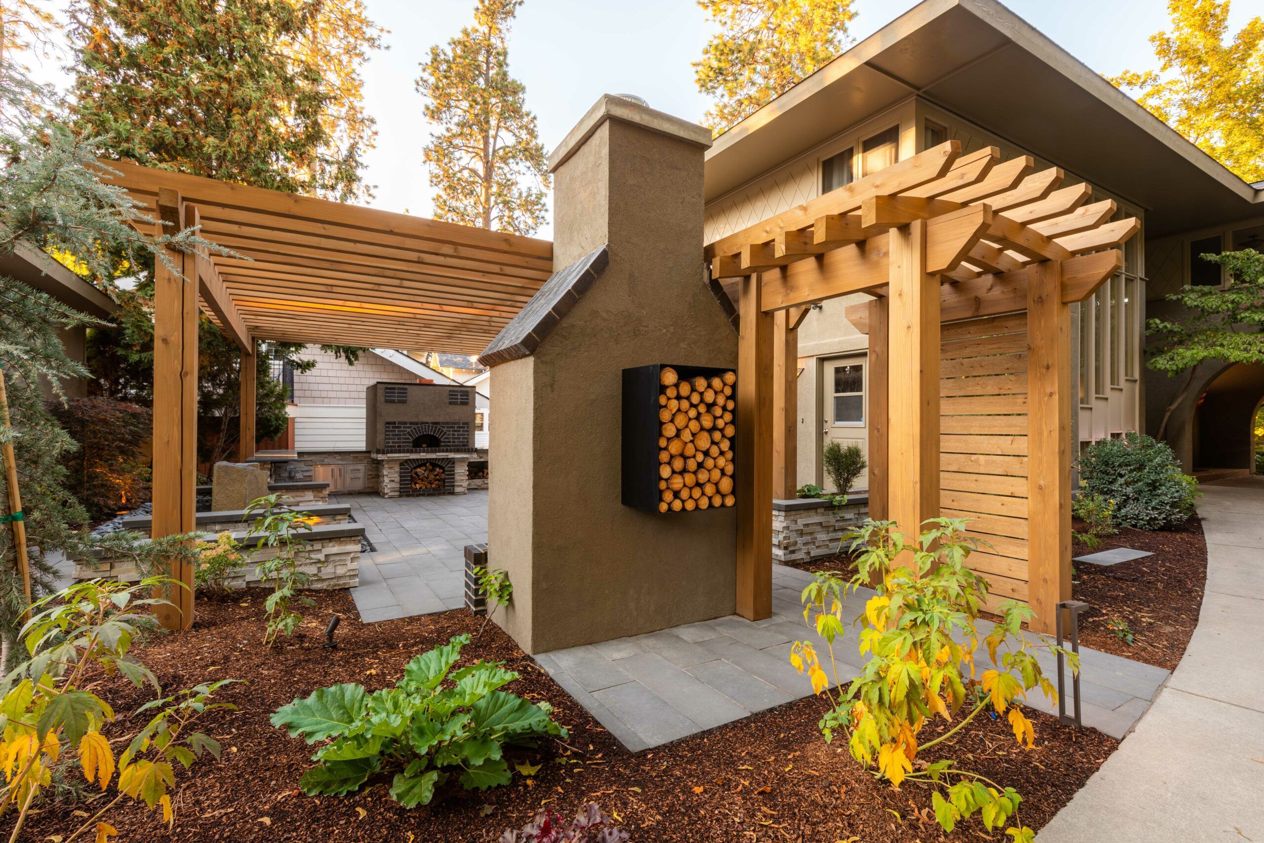 A modern outdoor patio area by Copper Creek Landscaping features wooden pergolas, a stack of firewood in a wall-mounted rack, garden beds with plants, and stone pavers. A curved sidewalk and a contemporary house are visible in the background.