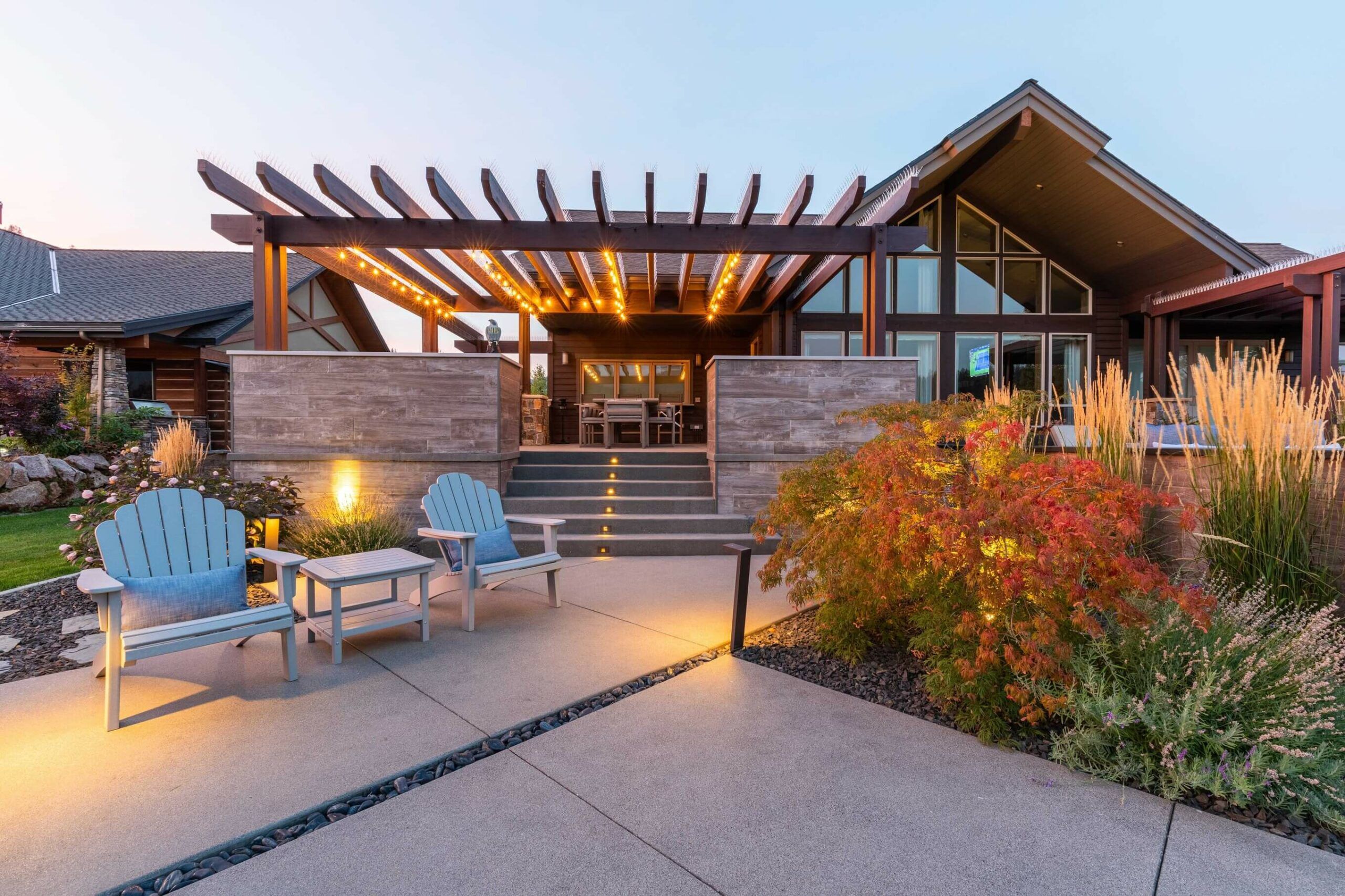 A modern house with a wooden pergola, string lights, outdoor seating, and expert landscaping by Copper Creek Landscaping features a concrete walkway and illuminated steps leading to the covered patio area, beautifully photographed at dusk.