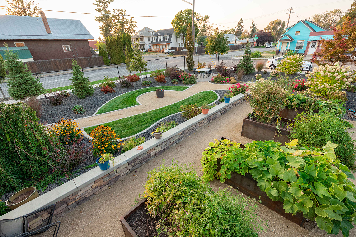 A landscaped garden by Copper Creek Landscaping features vegetable beds in the foreground, a curved gravel path, green lawn, assorted flowers, potted plants, trees, and houses in the background along a street.