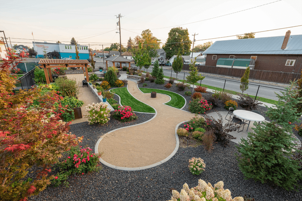 A landscaped garden by Copper Creek Landscaping features curved gravel paths, green grass, colorful flowering plants, a pergola, benches, and a round white table with chairs, all enclosed by a fence near a street and buildings.