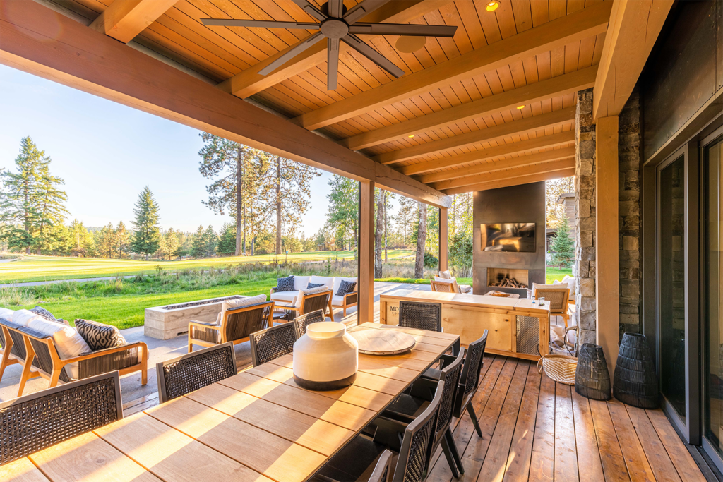 Covered outdoor patio with a wooden dining table, wicker chairs, ceiling fan, lounge area with sofas, fireplace, and TV. Large openings showcase lush green grass and trees—a beautiful example of Copper Creek Landscaping under a clear sky.