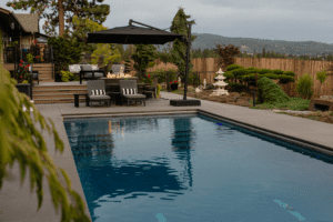 A modern backyard by Copper Creek Landscaping features a rectangular swimming pool, a patio with fire table and chairs under a large black umbrella, lush landscaping, and a wooden fence with hills visible in the background.