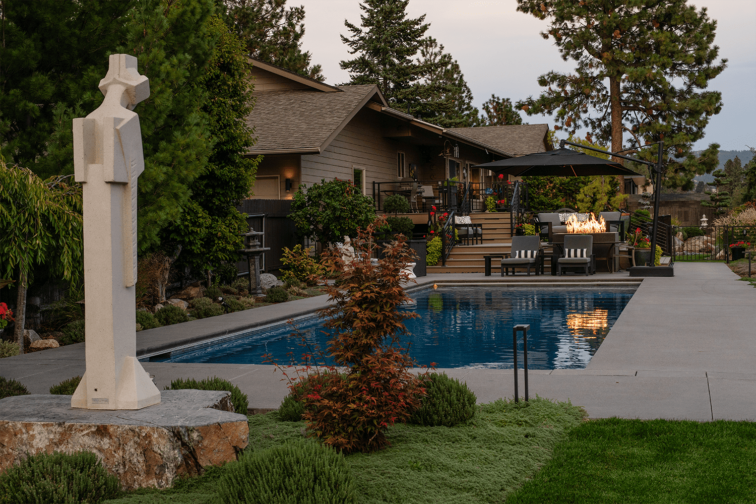 A backyard landscaped by Copper Creek Landscaping features a modern stone sculpture on a pedestal, a rectangular swimming pool, lounge chairs, fire pit, lush green shrubs, and a house with a wooden deck and patio area in the background.
