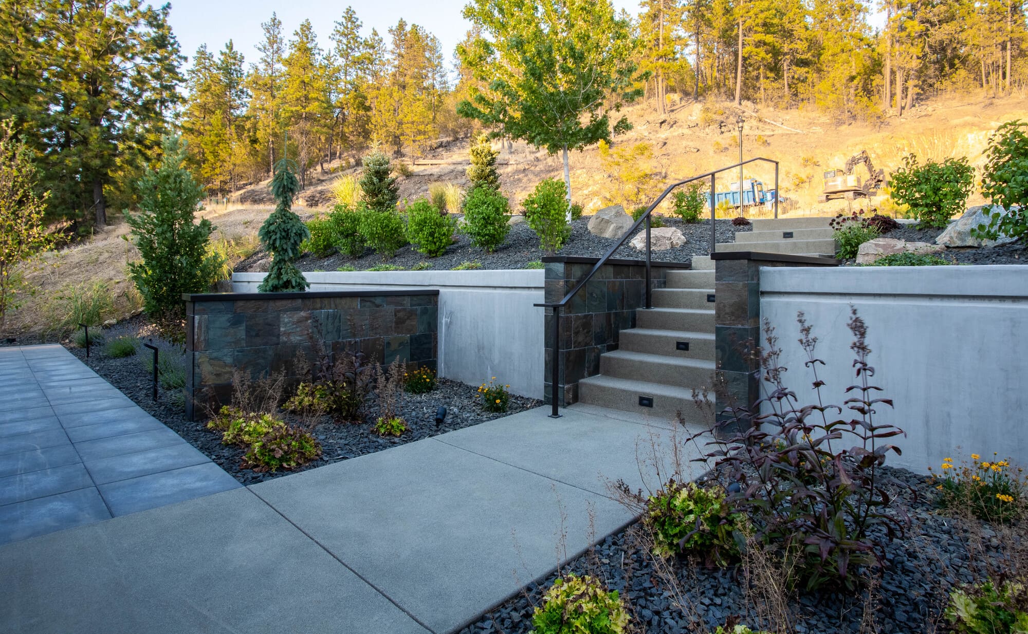 A beautifully landscaped outdoor area by Copper Creek Landscaping features concrete paths, steps with metal railings, stone-faced retaining walls, vibrant shrubs, and trees on a sunlit hillside.