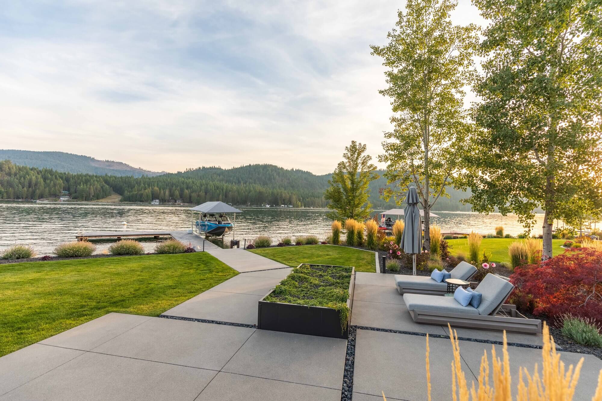 A modern lakeside patio with lounge chairs, manicured lawn, and a walkway—expertly crafted by Copper Creek Landscaping—leads to a dock. The scene overlooks a calm lake surrounded by forested hills under a partly cloudy sky.