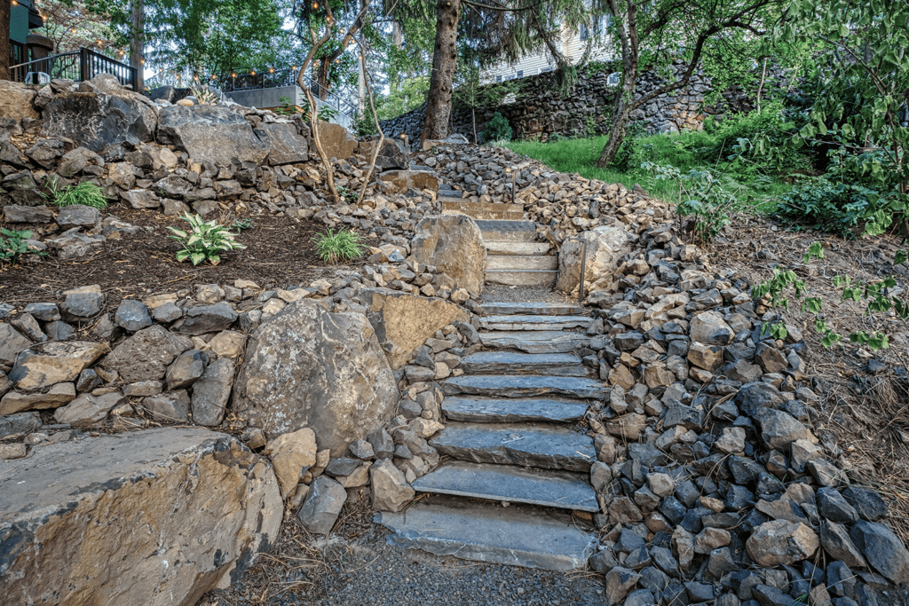 Stone steps lead up a landscaped slope by Copper Creek Landscaping, bordered by large and small rocks with green plants nestled among them. Trees and a rock wall complete the tranquil outdoor scene.