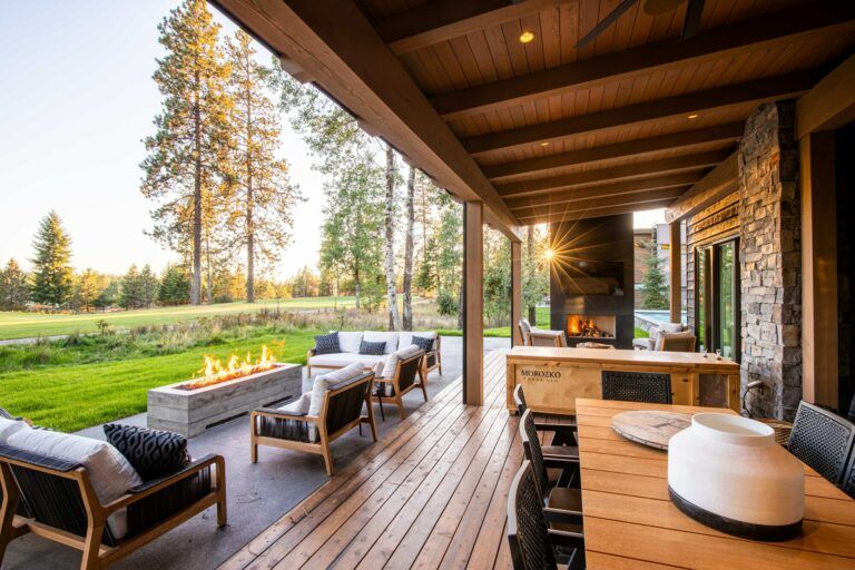 A covered wooden patio by Copper Creek Landscaping features a dining table, cushioned chairs, and an outdoor fireplace. Beyond the patio, lush landscaping includes green grass, tall trees, and a fire pit with seating, all lit by warm sunlight.