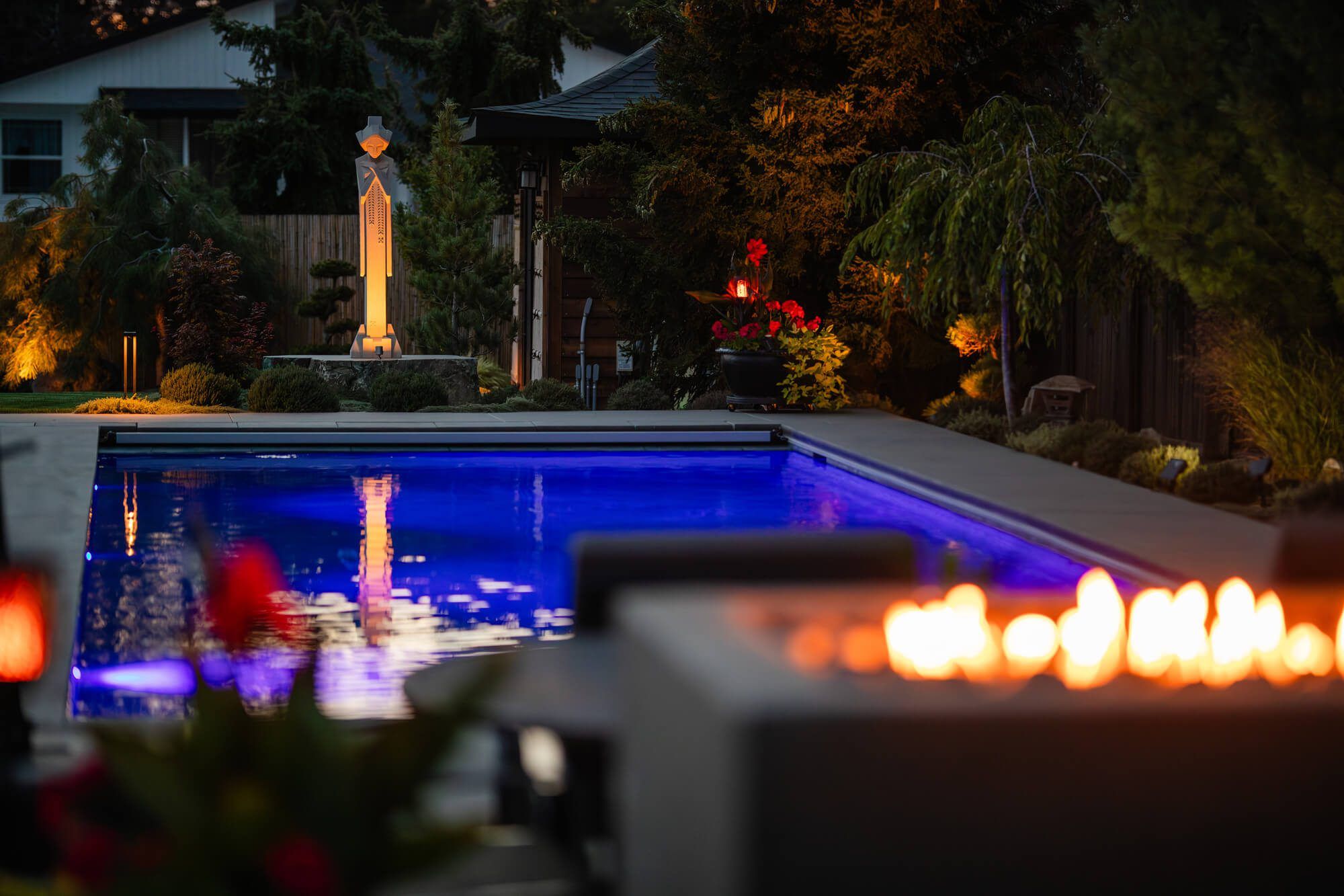 A backyard pool at dusk is lit with blue underwater lights. In the background, a statue is illuminated among trees and plants shaped by expert landscaping. In the foreground, a fire pit glows near modern outdoor furniture.
