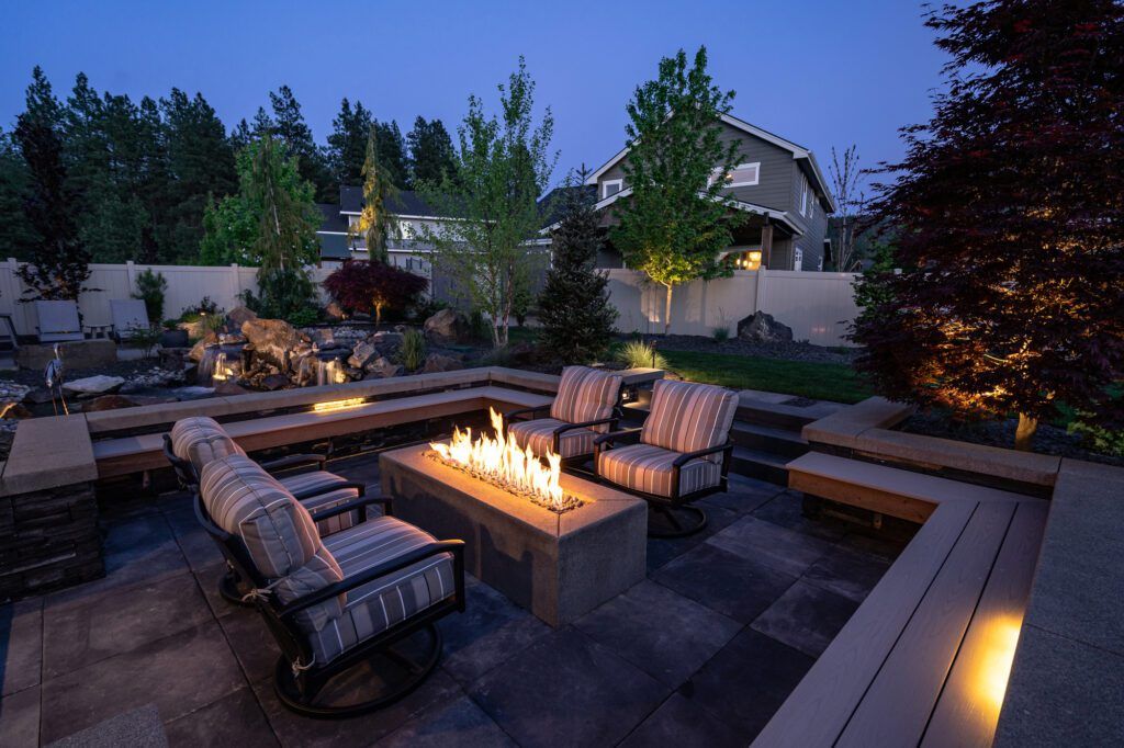 A backyard patio at dusk, crafted by Copper Creek Landscaping, features a rectangular fire pit surrounded by cushioned chairs and built-in benches, with elegant hardscaping, lush trees, and a cozy house in the background.