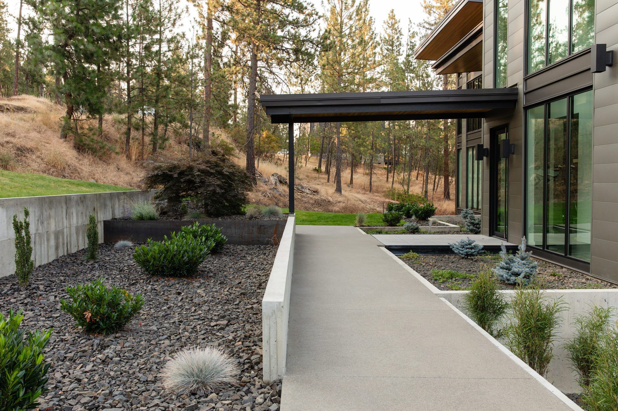 A modern house with large windows and a covered walkway, surrounded by minimalist Copper Creek Landscaping featuring rocks, shrubs, and concrete planters, set against a backdrop of trees and dry grass.