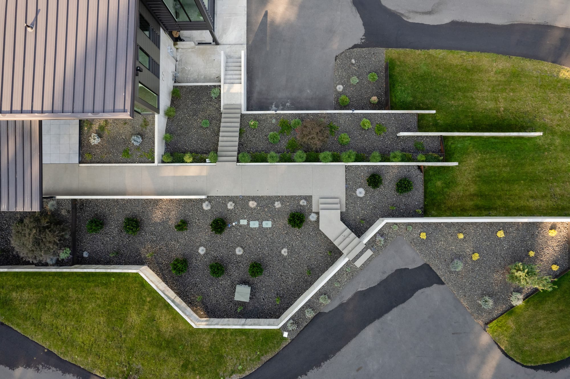 Aerial view of a modern building entrance with a concrete walkway, steps, and landscaped areas by Copper Creek Landscaping, featuring small shrubs and gravel, bordered by green grass and paved driveways.