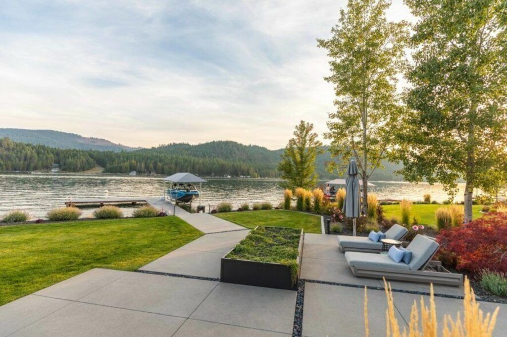 A landscaped patio by Copper Creek Landscaping with lounge chairs overlooks a calm lake and forested hills. A dock with a boat rests on the water, while trees and ornamental plants border the serene hardscaped area.