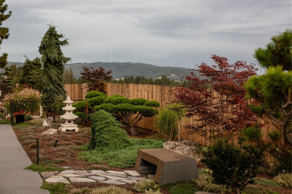 A landscaped Japanese-style garden by Copper Creek Landscaping features manicured trees, a stone path, a concrete bench, a stone lantern, and a bamboo fence, all set against rolling hills and a cloudy sky.