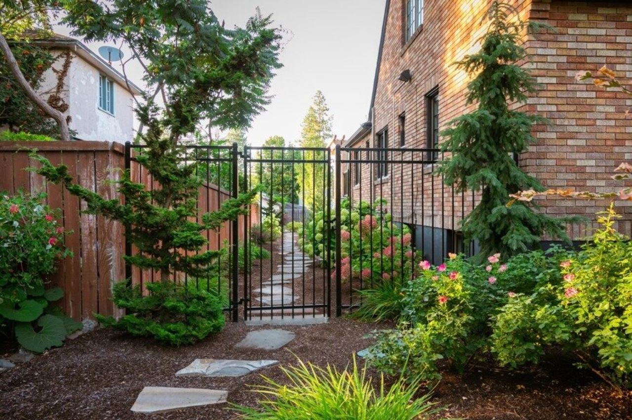 A black metal gate opens to a stone pathway lined with green shrubs and flowers, leading alongside a brick house. Tall trees and a wooden fence border the landscaped garden, showing expert landscaping by Copper Creek Landscaping.
