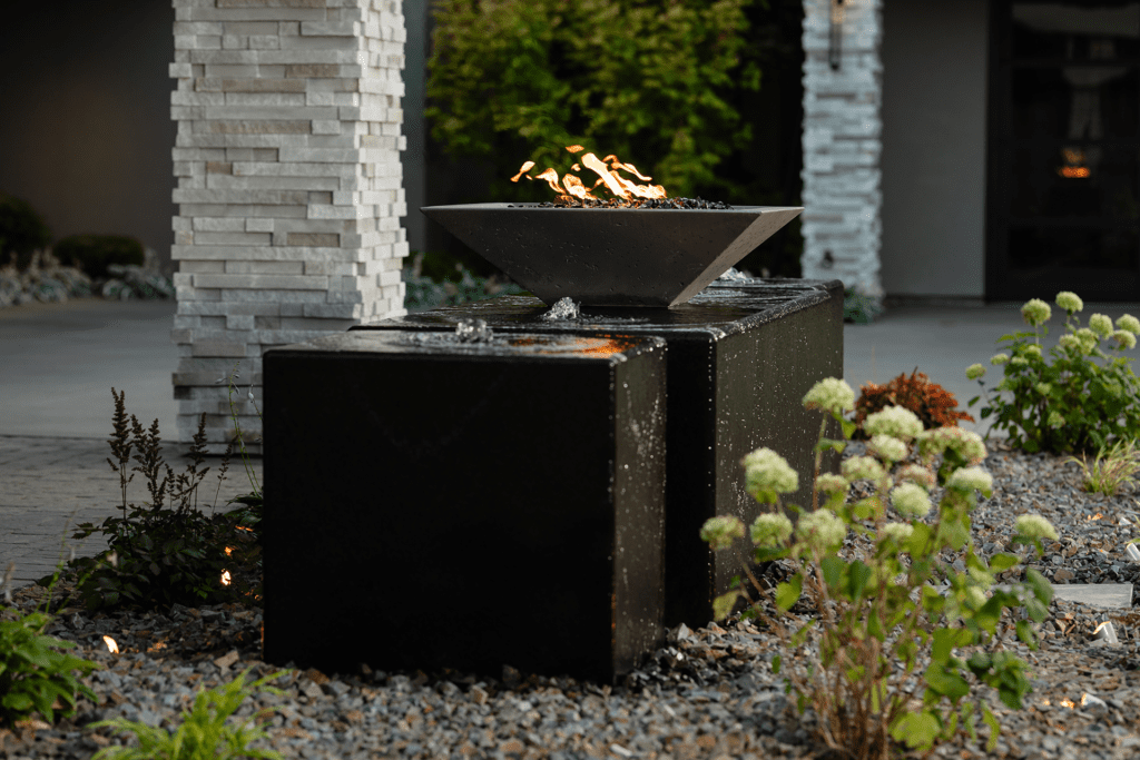 A modern outdoor water feature by Copper Creek Landscaping features a square black stone base, topped by a metal fire bowl with visible flames. Decorative plants and gravel surround the structure, with a stone pillar nearby.