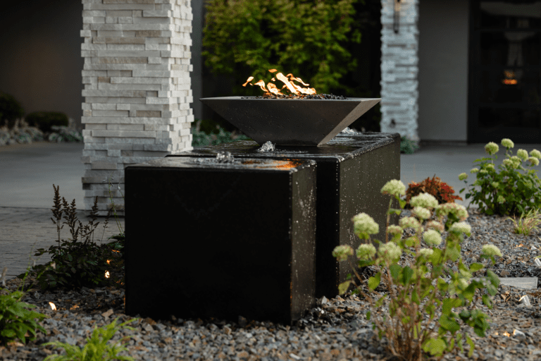 A modern outdoor water feature by Copper Creek Landscaping features a square black stone base, topped by a metal fire bowl with visible flames. Decorative plants and gravel surround the structure, with a stone pillar nearby.