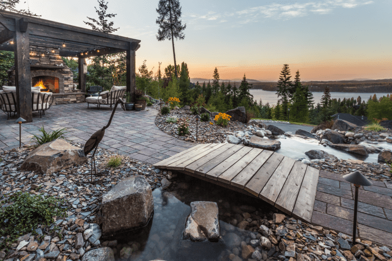 A landscaped backyard by Copper Creek Landscaping features a stone patio with outdoor seating, a fireplace, a wooden pergola with string lights, a small bridge over a stream, and views of the lake and trees at sunset.