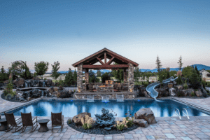 A rectangular swimming pool with a slide, surrounded by stone decking, outdoor seating, lush Copper Creek Landscaping, and a covered pavilion with stone columns and a fireplace in the background. Mountains are visible in the distance.