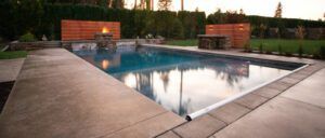 A rectangular outdoor swimming pool is surrounded by a concrete deck, with a firepit and seating area at one end and a stone bar or grill station on the side, all enhanced by Copper Creek Landscaping’s lush hedges and trees.
