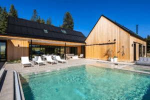 Modern house with light wood siding and black roof, large windows, and outdoor patio area. Professionally designed landscaping by Copper Creek Landscaping surrounds the swimming pool, lounge chairs, and adds beauty to the trees and blue sky in the background.
