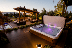 A modern backyard patio at dusk features a lit hot tub in the foreground, an outdoor seating area under a pergola with string lights, and a pool in the background surrounded by lush Copper Creek Landscaping.