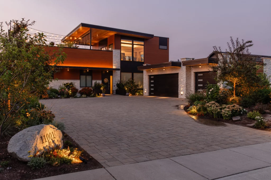 A modern two-story house with large windows, wood paneling, and stone accents. The brick driveway leads to two garages, while the front yard showcases Copper Creek Landscaping’s beautifully landscaped plants and illuminated outdoor lighting.