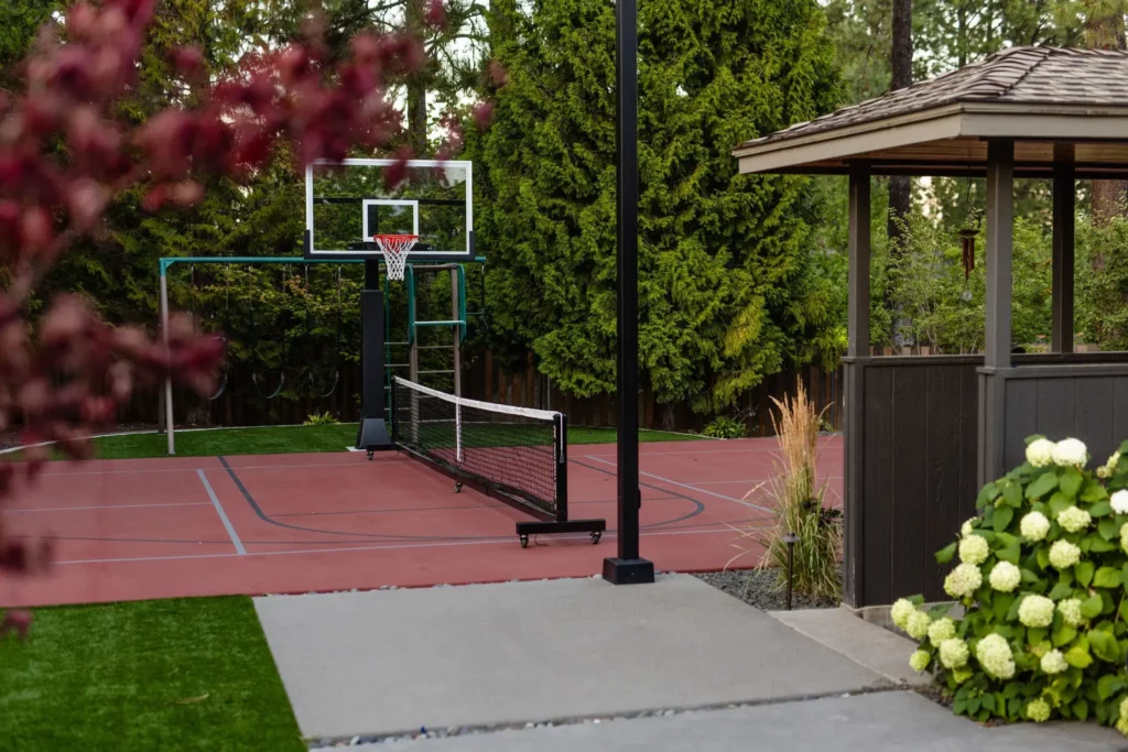 A backyard multi-use sports court by Copper Creek Landscaping features a basketball hoop and tennis net, surrounded by lush trees and greenery. A gazebo adorned with white hydrangeas is visible on the right side of the image.
