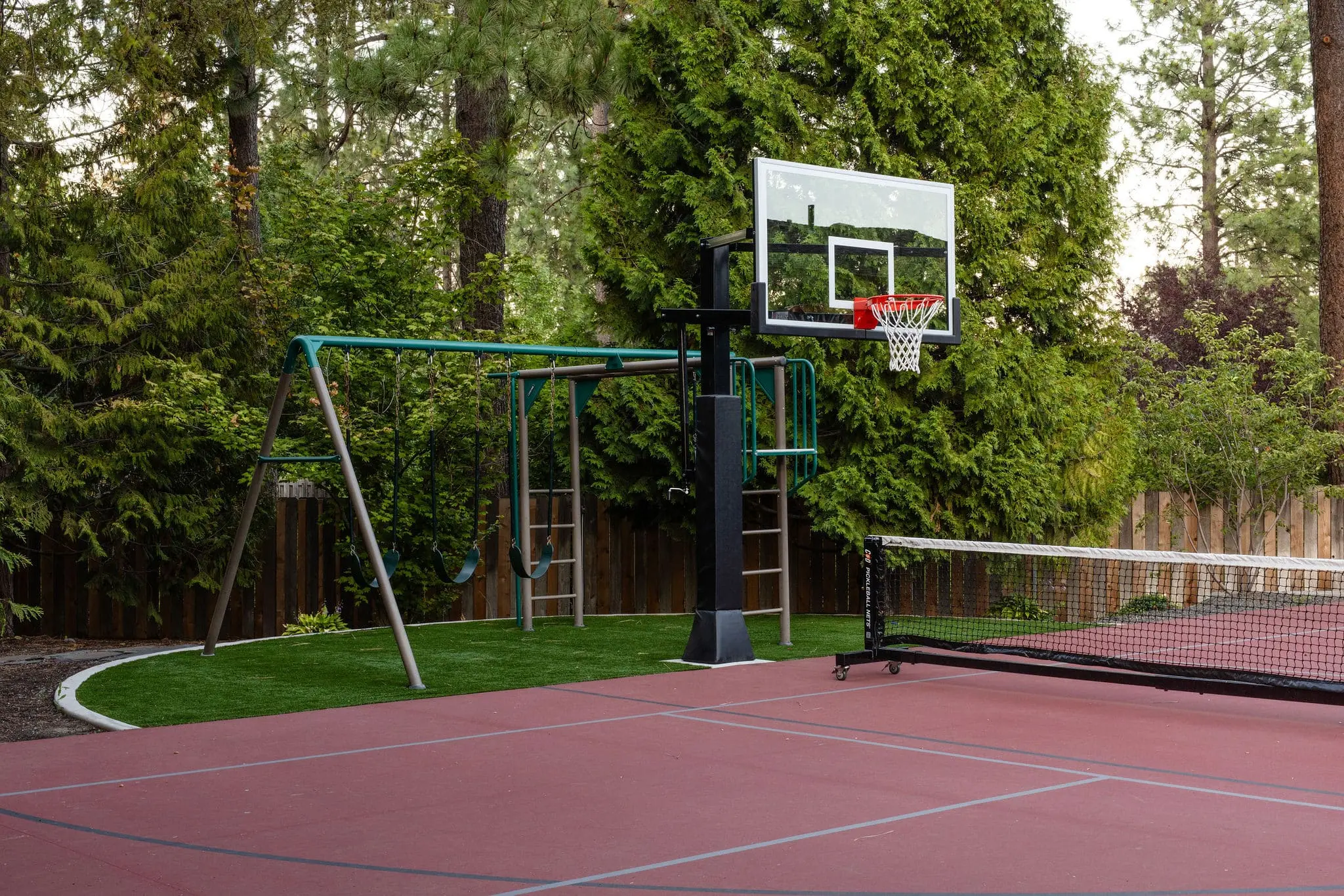 A red outdoor sports court by Copper Creek Landscaping features a basketball hoop, a swing set with two swings and a slide, and part of a tennis net, all surrounded by tall green trees and a wooden fence.