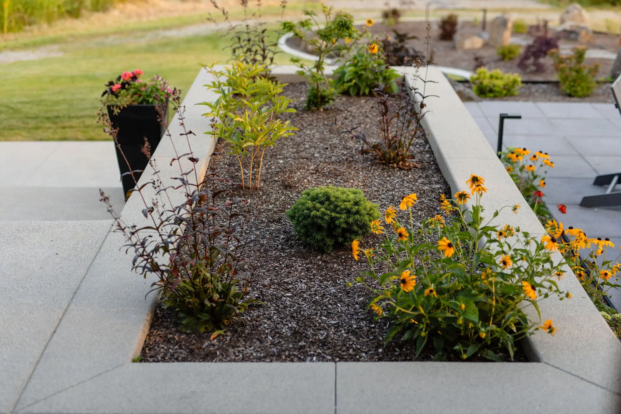 A rectangular concrete planter box from Copper Creek Landscaping contains green shrubs and yellow flowers, surrounded by soil. The planter sits on a patio with outdoor furniture and additional potted plants in the background.