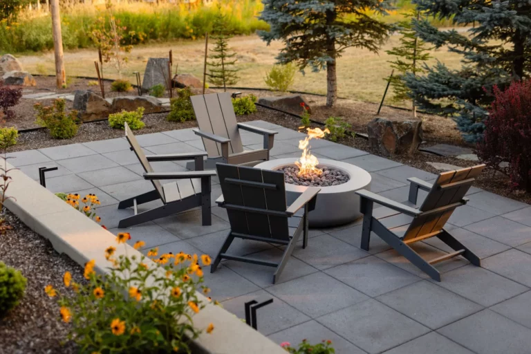 Four modern Adirondack chairs are arranged around a lit fire pit on a gray tiled patio. Thoughtful landscaping by Copper Creek Landscaping adds vibrant flowers and small shrubs, with pine trees and rocks creating a serene backdrop.
