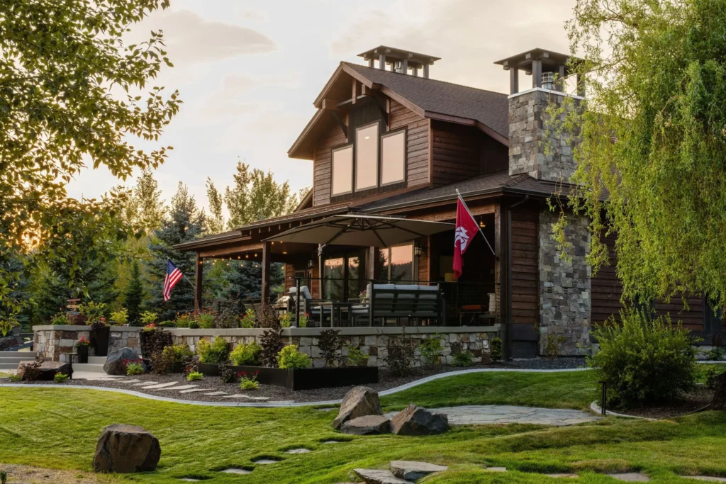 A two-story house with wood siding and stone accents, a covered porch with patio furniture, two flags, and a well-manicured lawn featuring Copper Creek Landscaping’s expertly designed trees and landscaped garden beds in the foreground.
