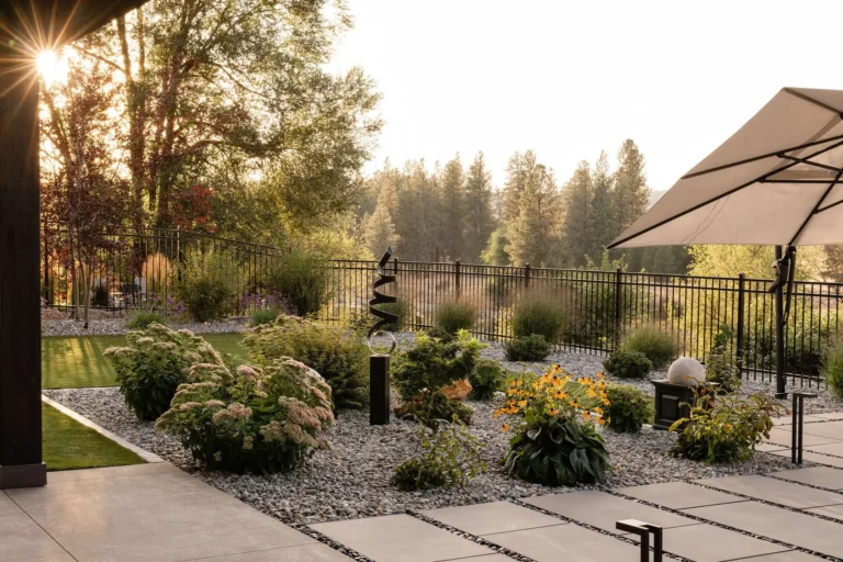 A landscaped backyard by Copper Creek Landscaping features stone pathways, decorative gravel, and garden beds with shrubs and flowers. A black metal fence runs along the back, with an umbrella for shade and sunlight filtering through the trees.