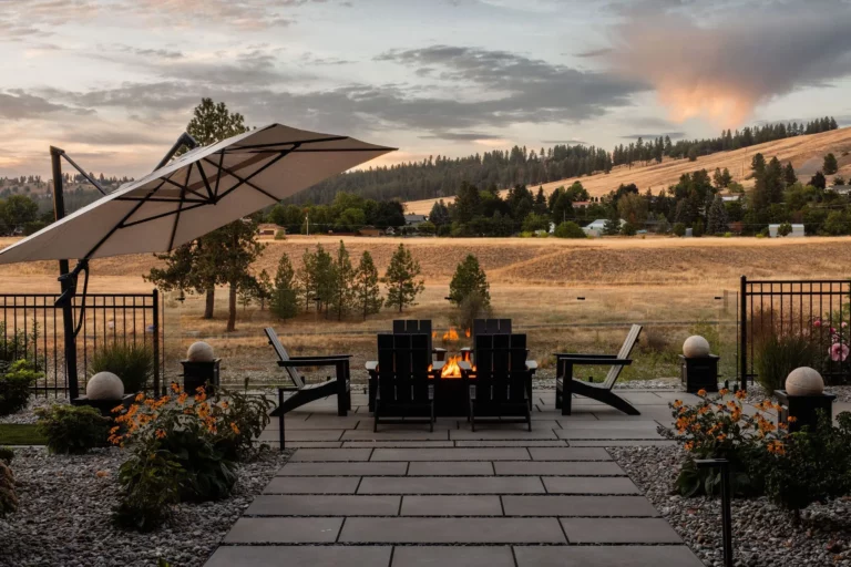 A patio with four black chairs around a fire pit, under a large umbrella, overlooks a rural landscape at sunset. Orange flowers and stone paths by Copper Creek Landscaping enhance the view of fields, trees, and hills.
