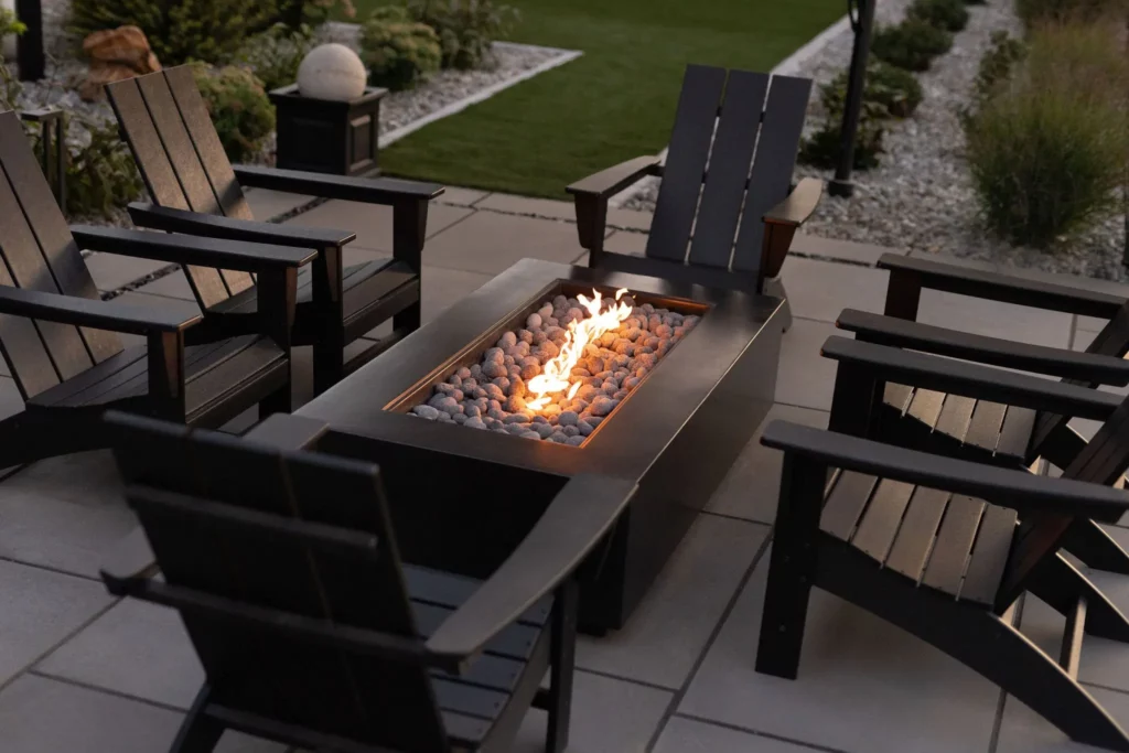 Rectangular fire pit with stones and flames at the center of a patio, surrounded by six black Adirondack chairs on stone tiles, with lush grass and Copper Creek Landscaping’s expert landscaping in the background.