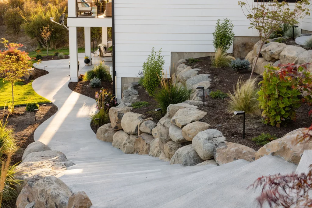 Concrete steps bordered by large rocks and landscaped plants lead up to a white house. Copper Creek Landscaping designed a curved concrete path that winds alongside the garden, featuring shrubs, small trees, and fresh mulch.