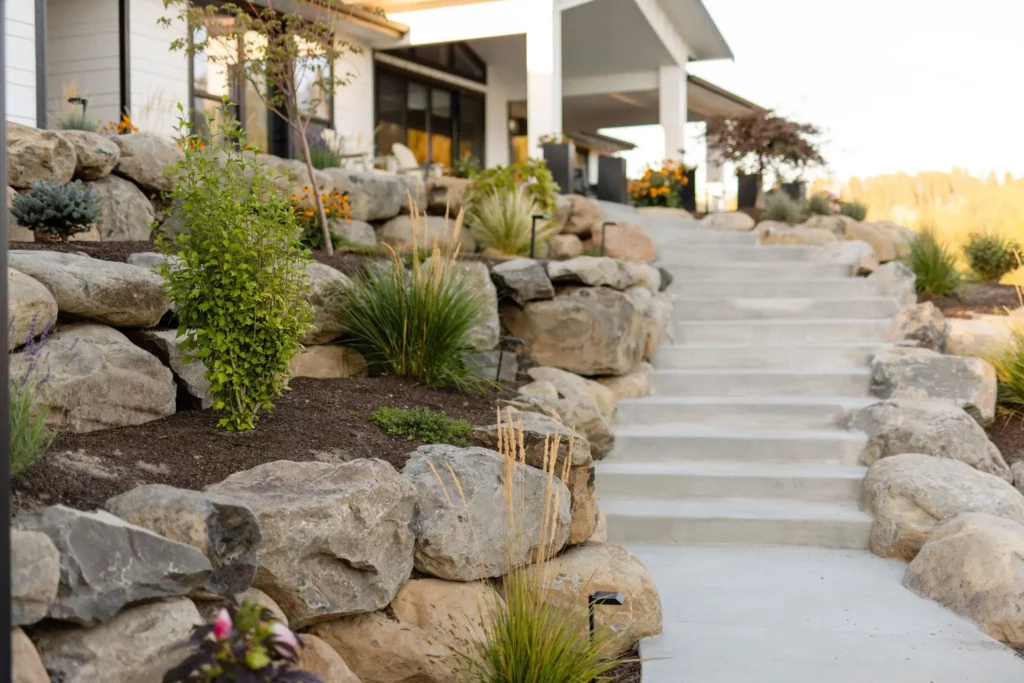 A concrete staircase bordered by large rocks and landscaped plants leads up to a modern house with white siding and a covered porch, showcasing expert Landscaping by Copper Creek Landscaping. Shrubs, grasses, and flowers flourish among the rocks.