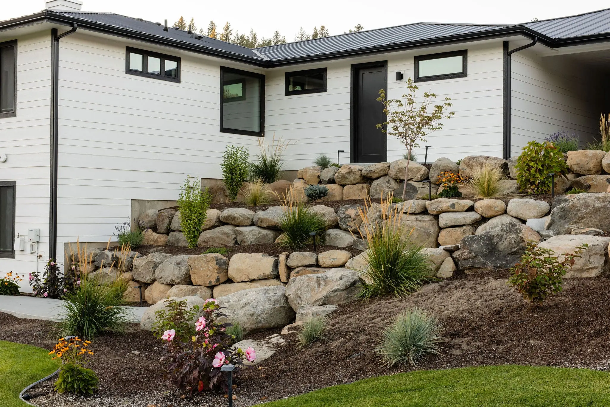 A modern white house with black trim sits on a slope. The landscaped front yard by Copper Creek Landscaping features large stone retaining walls, rocks, various shrubs, flowers, and mulch, with green grass in the foreground.