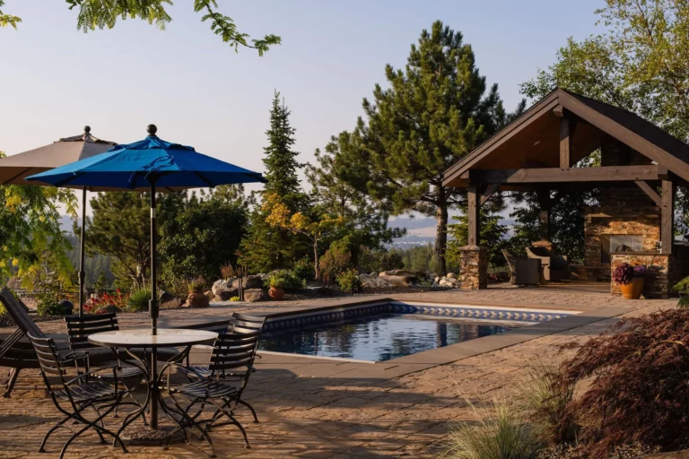 A backyard patio by Copper Creek Landscaping features a small rectangular pool, metal table with chairs under a blue umbrella, and a covered outdoor fireplace. Trees and plants surround the area, with a scenic view in the background.