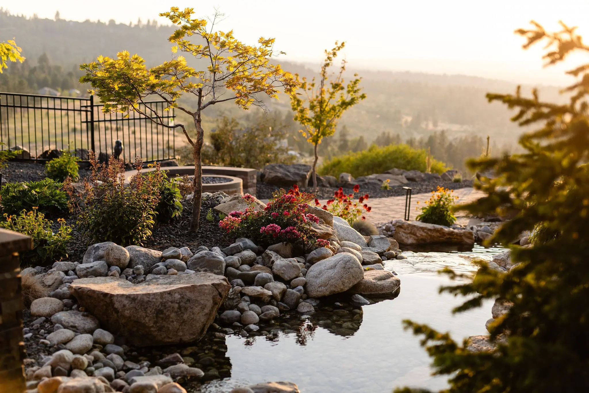 A landscaped garden by Copper Creek Landscaping features a small pond surrounded by rocks, shrubs, and flowering plants. Trees and a metal fence line the background, with hills visible in the hazy distance at sunset.