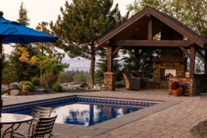 A backyard scene designed by Copper Creek Landscaping features a rectangular swimming pool, stone patio, metal chairs and table, an outdoor fireplace under a wooden pavilion, and lush trees and plants surrounding the area.