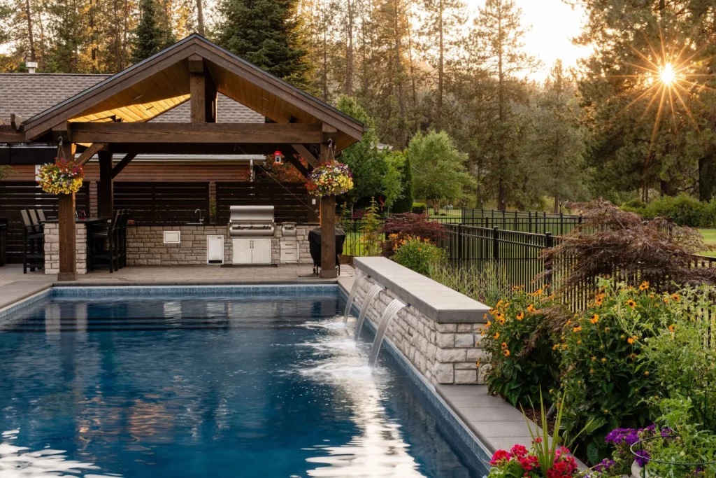 A backyard pool with a stone waterfall feature, designed by Copper Creek Landscaping, is surrounded by flowers and greenery. An outdoor kitchen under a wooden pergola, black metal fence, and trees complete this beautiful sunset scene.