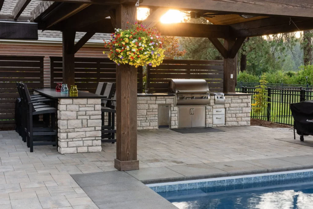 Outdoor kitchen and dining area under a wooden pergola by a pool, featuring a stainless steel grill, stone counters, bar seating, hanging flower basket, and privacy fence with sunlight filtering through trees by Copper Creek Landscaping.