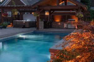 A backyard by Copper Creek Landscaping features a lit swimming pool, a covered patio with outdoor kitchen, hanging flower baskets, and a hot tub in the background, with warm lighting and expert landscaping around a small tree in the foreground.