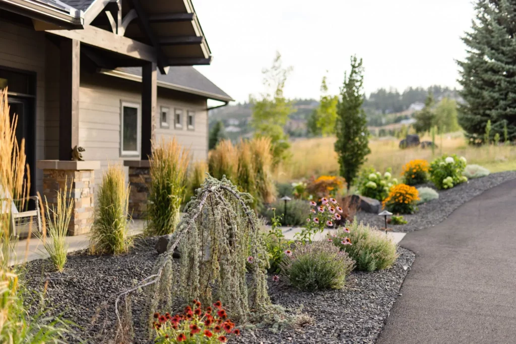 A landscaped front yard by Copper Creek Landscaping features drought-tolerant plants, ornamental grasses, and flowering perennials beside a paved driveway and a house with stone and wood siding, set against open grassy land and trees.