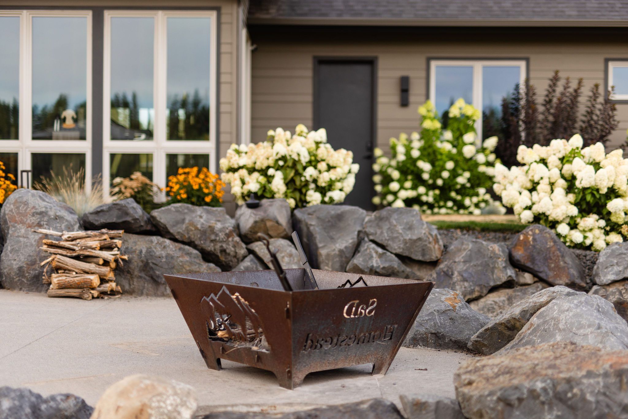 A square metal fire pit with mountain designs sits on a stone patio, beautifully integrated by Copper Creek Landscaping, surrounded by large rocks, blooming white and yellow flowers, and a small stack of firewood in front of a house with large windows.