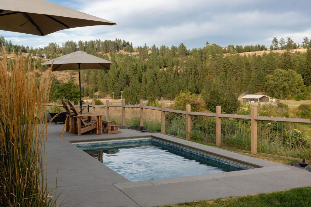 A small rectangular pool is surrounded by a concrete patio with wooden lounge chairs and umbrellas. Thoughtful landscaping by Copper Creek Landscaping enhances the area, bordered by a wooden fence, with trees and hills under a partly cloudy sky.