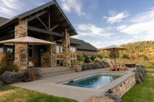 A modern house with a stone and wood terrace overlooks a small rectangular pool, surrounded by a concrete deck, patio chairs, and umbrellas. The lush grassy yard with hills and trees showcases expert Landscaping by Copper Creek Landscaping.